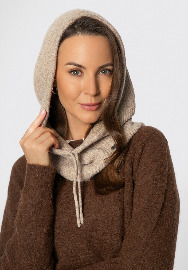 Beige hoodie van een wolmix met een geribbelde structuur en trekkoorddetail. Het model draagt het gelaagd over een donkerbruine trui, waardoor het ontwerp goed zichtbaar is.