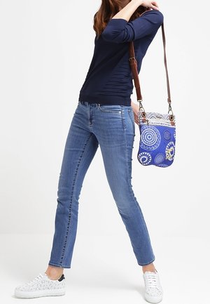 Granatowa bluzka z długim rękawem, jasnoniebieskie dopasowane dżinsy, białe trampki oraz niebieska torebka w wzory z brązowym paskiem, z okrągłymi motywami.