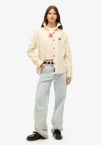 Giovane donna che indossa una camicia in velluto a coste beige sopra una maglietta grafica rosa chiaro, jeans larghi azzurri, scarpe nere e una cintura nera.