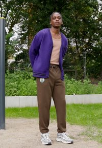 Cardigan viola sopra un top beige chiaro, pantaloni joggers marroni e sneakers grigie. Design casual con chiusura a bottoni e logo sui pantaloni.