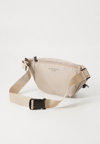 Beige nylon waist bag with a textured finish, adjustable strap, black hardware, and a front zip compartment. Features logo print.