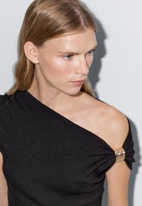 Svart axelbandslös topp med en vriden design, prydd med en gyllene metallaccent vid axeln. Slät textur och åtsittande silhuett.