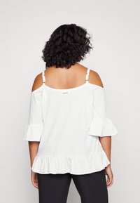 Top bianco scivolato sulle spalle con maniche e orlo arricciati. Tessuto testurizzato con spalline regolabili e un piccolo logo sul retro.