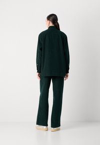 Set di camicia e pantaloni in corduroy verde scuro, con una vestibilità comoda, polsini abbottonati e tessuto testurizzato, abbinato a scarpe di colore chiaro.