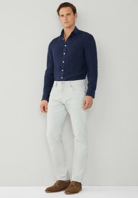 Camisa de lino azul marino de botones, jeans ajustados gris claro y zapatos de ante marrón. Hombre de pie con las manos en los bolsillos contra un fondo neutro.
