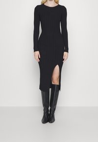 Abito a maglia nera a coste con maniche lunghe, di lunghezza media, con uno spacco alto fino coscia, abbinato a stivali in pelle nera alti fino al ginocchio.
