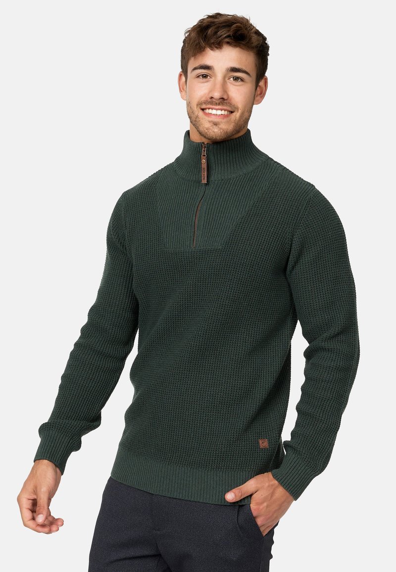 Suéter de punto verde oscuro con un cuello alto y media cremallera. Puños y dobladillo acanalados, con un panel frontal texturizado y un sutil detalle de logotipo.