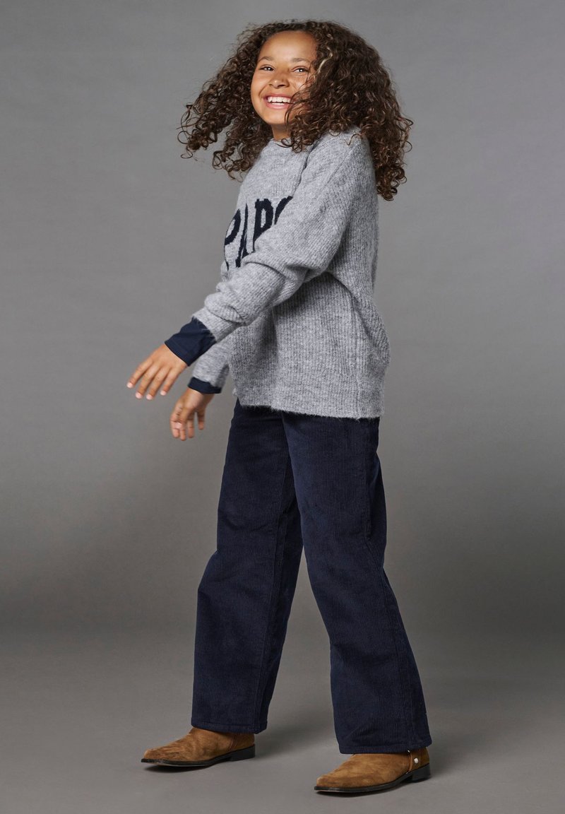 Pull en tricot gris avec le mot "PARIS" en bleu marine, associé à un pantalon en velours côtelé bleu marine et des bottines marron. L'enfant semble joyeux, en mouvement.