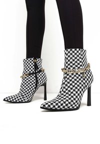 Černobílé kotníkové boty s vzorem houndstooth, s špičatou špicí, vysokými stiletto podpatky, zlatými řetízkovými akcenty a předním zipem.