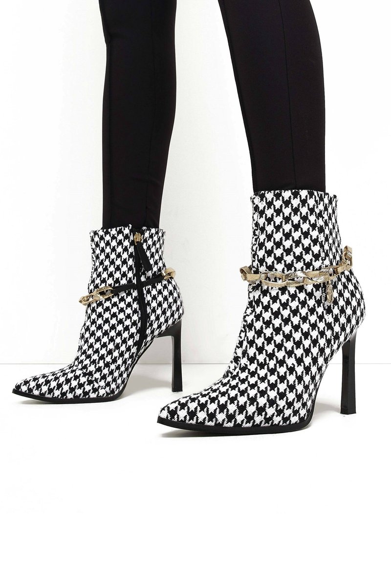 Černobílé kotníkové boty s vzorem houndstooth, s špičatou špicí, vysokými stiletto podpatky, zlatými řetízkovými akcenty a předním zipem.