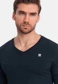 Maglietta a maniche lunghe blu navy con scollo a V realizzata in tessuto morbido, dotata di un piccolo logo bianco sul lato sinistro del petto. Texture liscia con design minimale.