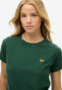 Blonde vrouw met los haar, gekleed in een donkergroen Superdry t-shirt, kijkt met een neutrale uitdrukking naar de camera.