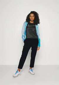 Nike-Windbreaker in blauem Farbverlauf mit langen Ärmeln, schwarzem Sportoberteil, schwarzen schmal zulaufenden Hosen und wei�ßen Sneakers mit blauen und rosa Akzenten.