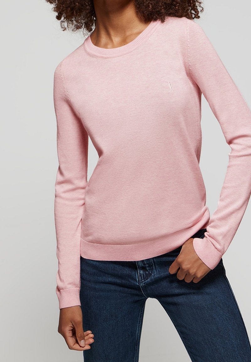 Femme portant un pull à manches longues rose clair et un jean bleu foncé, une main dans la poche, sur un fond uni.
