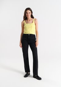 Geel gebreide tanktop met dikke bandjes en bloemige versieringen, gecombineerd met hoge zwarte denim jeans en zwarte schoenen.