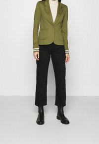 Blazer fitted verde oliva con due tasche frontali, dettagli a righe bianche sulle maniche, abbinato a pantaloni neri a gamba dritta e stivaletti neri.