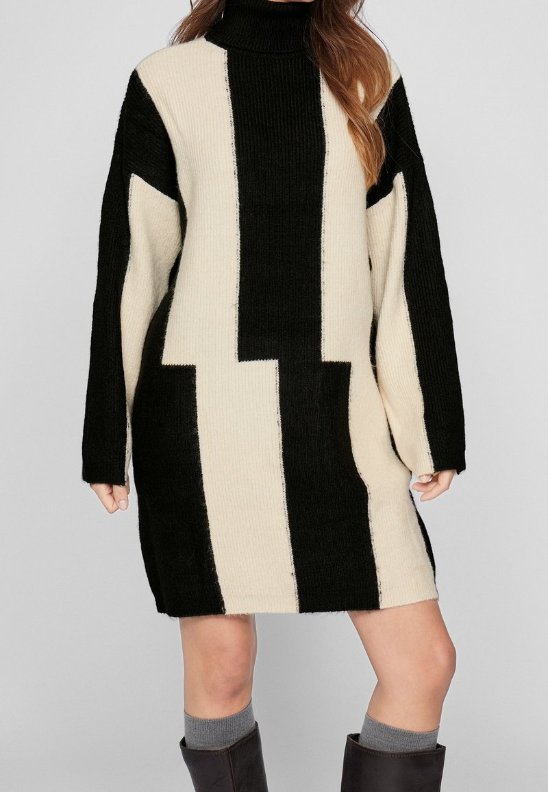 Robe-pull tricotée à col haut, présentant des blocs de couleur audacieux noir et crème, des manches surdimensionnées et un motif côtelé texturé.
