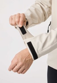 Roka nastavlja bež manšeto jakne s črnim ježkom na svetli koži na belem ozadju.