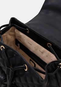 Sac à main noir matelassé avec quincaillerie dorée. Présente un intérieur beige avec un motif "GUESS", une poche zippée et une fermeture à cordon.
