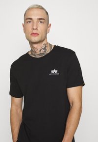 Svart bomull T-skjorte med rund hals, korte ermer og hvit "Alpha Industries" logo på brystet. Glatt tekstur.