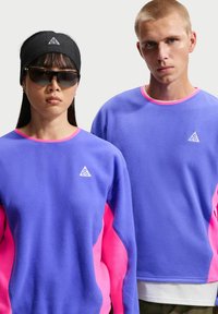 Δύο μοντέλα φορούν ταιριαστές μπλούζες σε μπλε-μοβ και ροζ χρώμα με λογότυπα ACG· το ένα φορά γυαλιά ηλίου και μαύρη κορδέλα στο κεφάλι.