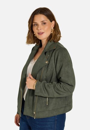 Frau mit kurzen braunen Haaren, die goldene Creolen trägt, eine olivgrüne Jacke über einem weißen Oberteil und blaue Jeans, schaut leicht nach rechts.