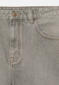 Gros plan sur un jean en denim gris clair montrant la taille, le bouton, la boucle de ceinture, la poche avant et les détails de couture.