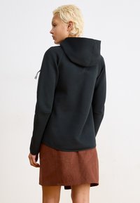 Sweatshirt à capuche noir avec une texture lisse, doté d'un cordon de serrage, associé à une jupe marron. Le design est simple et moderne.