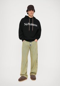 Sudadera negra con la impresión "No Problemo", pantalones de pierna ancha beige a rayas y zapatos marrones sin cordones con detalles bordados. Gorro marrón.