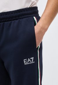 Κοντινό πλάνο ενός ατόμου που φοράει σκούρο μπλε φόρμα EA7 Emporio Armani με το χέρι στην τσέπη και ριγωτή ταινία με το λογότυπο κατά μήκος της πλαϊνής ραφής.