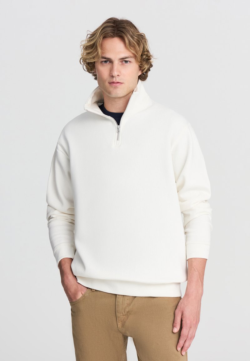 Pullover bianco in pile con mezzo zip, polsini e orlo a costine. Indossato sopra una camicia blu navy, abbinato a pantaloni color khaki. Tessuto morbido.