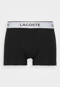 Black cotton boxer briefs with a white elastic waistband featuring the "LACOSTE" logo. Simple design, mid-thigh length, soft texture.