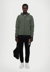 Felpa con zip verde con logo, abbinata a pantaloni della tuta neri con una stampa bianca. Il modello tiene una borsa a tracolla nera e indossa sneakers bianche.