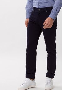 Pantaloni di cotone blu navy con taglio slim, dotati di chiusura a bottone e risvolti, abbinati a sneaker bianche texturizzate.