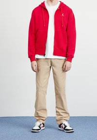 Persoon in een rode zip-up hoodie, witte T-shirt, beige broek en zwart, wit en rode sneakers die op een blauw tapijt staat.