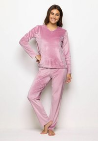 Donna che sorride, in piedi a piedi nudi, indossando un morbido top lungo in velluto rosa chiaro e pantaloni abbinati, su uno sfondo bianco semplice.