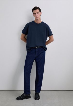 Tricou bleumarin cu mâneci scurte, combinat cu pantaloni din denim închis la culoare și pantofi negri. Design simplu și croială lejeră, cu decorațiuni minimale.