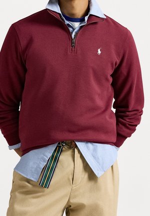 Mężczyzna ubrany w bordowy sweter na zamek do połowy wysokości, na jasnoniebieską koszulę zapinaną na guziki, beżowe spodnie oraz pasiasty materiałowy pasek.