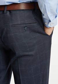 Pantaloni a scacchi blu scuro con una texture liscia, dotati di una tasca posteriore con bottone e una cintura marrone, abbinati a una camicia azzurra.