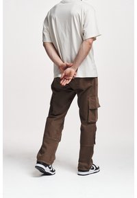 Beige bomulls-T-shirt och bruna cargobyxor med fackfickor; svarta och vita sneakers; visad bakifrån i en neutral miljö.