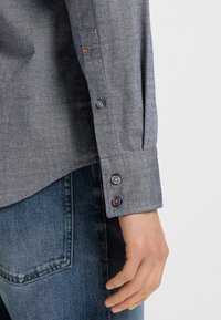 Grijze button-up overhemd met een gestructureerd patroon, met een enkele oranje stiksel op de mouw. Draagt met blauwe denim jeans.