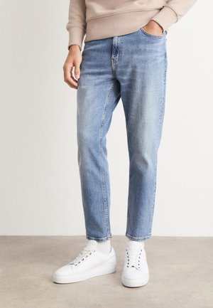 Jeans de mezclilla azul claro con corte recto, diseño de cinco bolsillos y textura desgastada, combinados con zapatillas blancas y un sudadera beige.