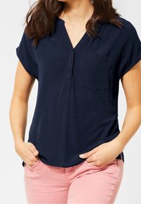 Navyblauwe blouse met korte mouwen en een V-hals, voorzien van een borstzakje; zachte stof, ruime pasvorm, gecombineerd met lichtroze broek.