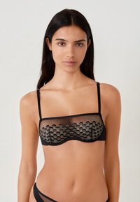 Bralette in rete nera con pannello trasparente decorato da motivi circolari testurizzati, spalline regolabili e fascia liscia sotto.