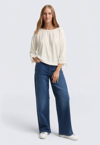 Ecrufarbenes Longsleeve-Top mit rundem Ausschnitt und Bindebändern an den Ärmeln, kombiniert mit weit geschnittenen blauen Jeans und weißen Turnschuhen.