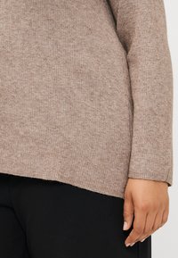 Light brown ribbed knit sweater with a slightly longer back hem and fitted sleeves, paired with black trousers. Visible texture details.