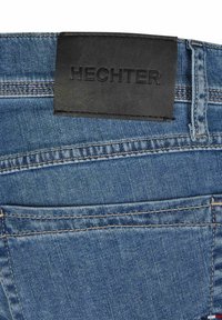 Denim jeans met een donkerleren merklabel met de tekst "HECHTER," blauwe stof met opvallende stiksels en een klassieke vijf-pocketontwerp.
