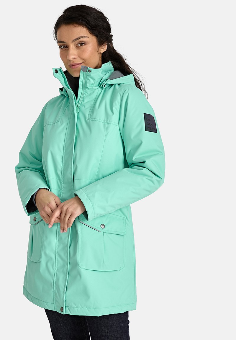 Mėtų žalios spalvos nepralaidi vandeniui striukė su užtrauktuku, reguliuojama kapišonu, dviem priekinėmis kišenėmis ir juodu logotipu ant kairiojo rankovės.