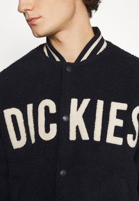 Giacca in pile nera con superficie texturizzata, dotata di un logo color crema, chiusura con bottoni e colletto a righe in contrasto.