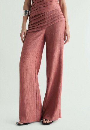 Mujer con pantalones amplios de talle alto en tono rosa polvo, con top texturizado plisado y pulsera plateada, de pie frente a un fondo liso.